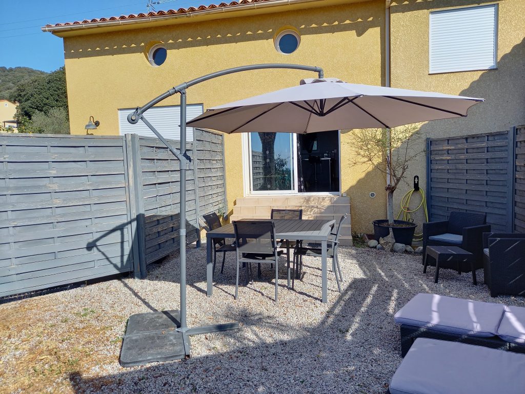 Photo qui représente l'espace jardin privatif de Mèrula, composé d'une table de jardin, d'un salon et deux transats avec un parasol et d'une plancha